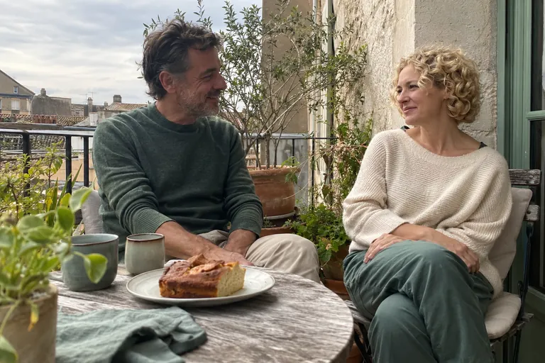 Deux adultes discutent assis à une table de balcon avec un gâteau et deux tasses.