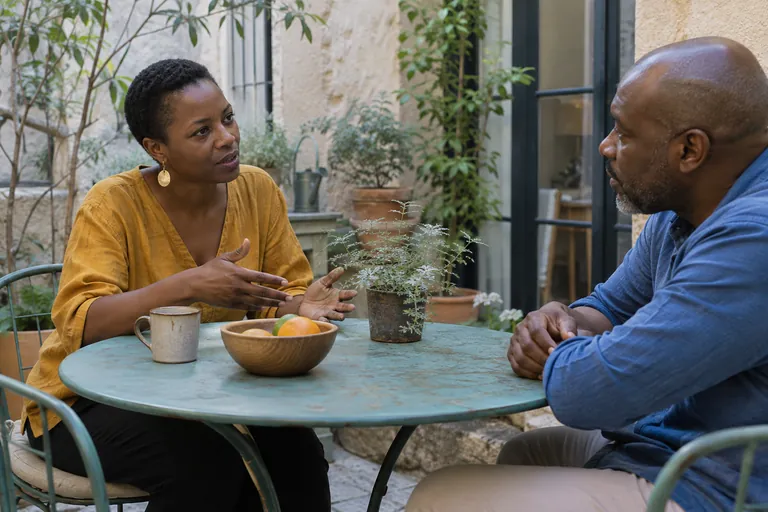 Deux adultes discutent à une table extérieure avec un bol de fruits.