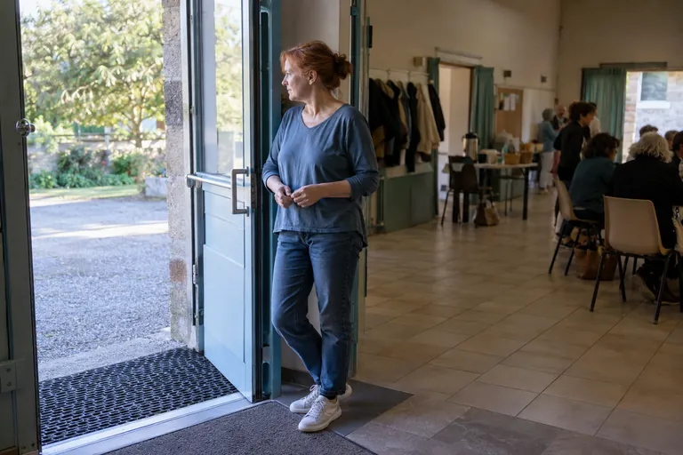 Une femme attend près de la porte ouverte d’une salle où plusieurs personnes sont réunies.