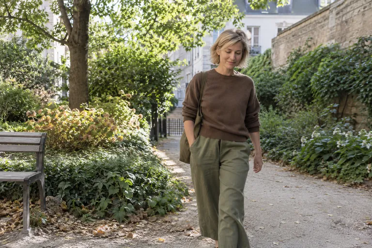 Une femme marche sur une allée arborée avec un sac à l’épaule.