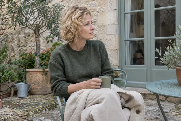 Une femme assise dans une cour tient une tasse et regarde sur le côté.