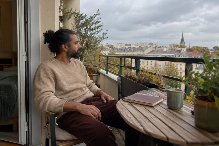 Un homme assis sur un balcon regarde dehors, un carnet fermé sur la table.