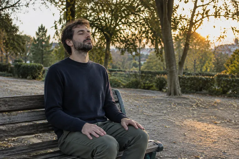 Un homme assis sur un banc de parc au coucher du soleil respire calmement, les mains posées sur les genoux