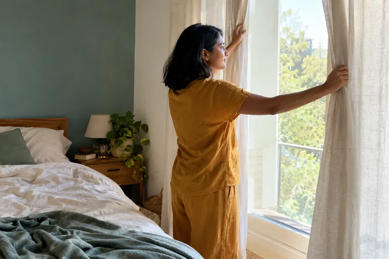 Une femme ouvre les rideaux d’une chambre éclairée par le soleil.