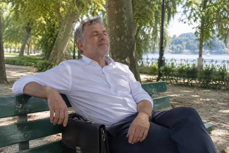 Un homme assis sur un banc de parc sous les arbres, les yeux fermés, sa sacoche posée à côté de lui