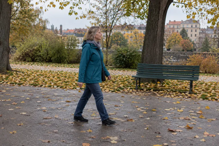 Une femme en veste marche sur une allée de parc couverte de feuilles d’automne.