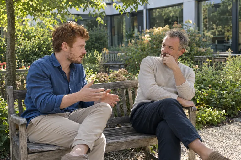 Deux hommes discutent assis sur un banc dans un jardin.