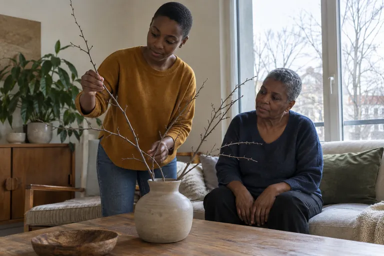 Une femme dispose des branches dans un vase tandis qu’une autre la regarde depuis un canapé.