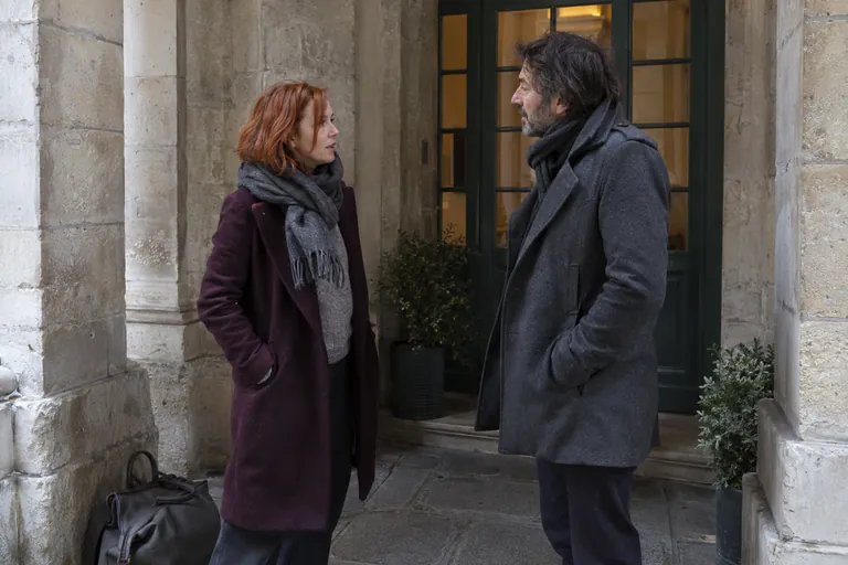 Deux adultes en manteau discutent sous un porche.