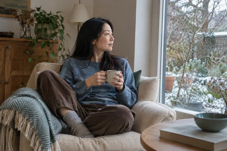 Une femme assise dans un fauteuil tient une tasse et regarde par la fenêtre.