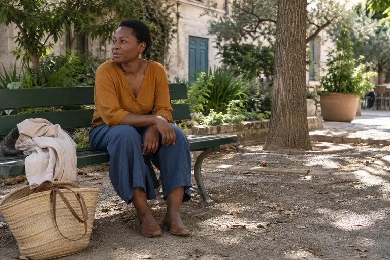 Une femme assise sur un banc regarde une place, un panier posé au sol à côté d’elle.