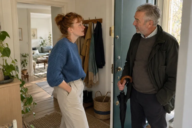 Deux personnes discutent sur le seuil, l’homme tenant un parapluie fermé.