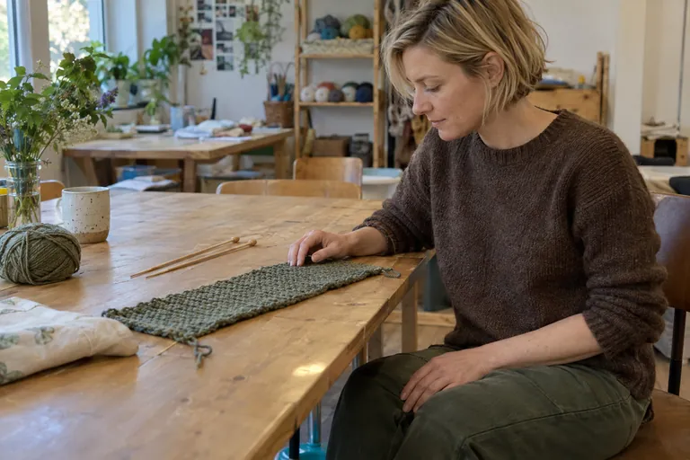 Une femme regarde un ouvrage en laine posé sur une table, les aiguilles à côté.